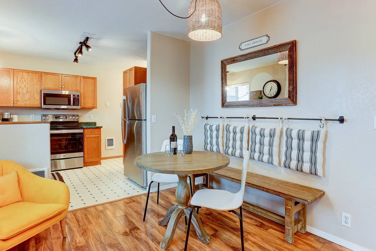 This image features a round wooden table with a yellow accent chair and striped bench seating, set near kitchen appliances. The scene suggests a space ideal for casual dining, enjoying coffee, or relaxing with a book, surrounded by a relaxed and inviting ambiance.