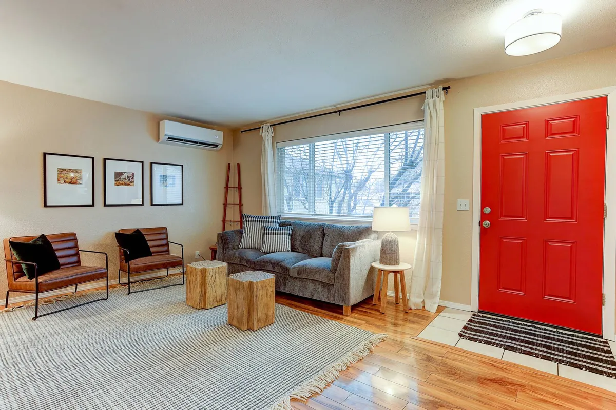 A bright living area anchored by a large window that lets in abundant natural light, a comfortable seating arrangement, a wooden coffee table, and the iconic red front door. The scene radiates coziness, making it perfect for relaxing or entertaining guests.