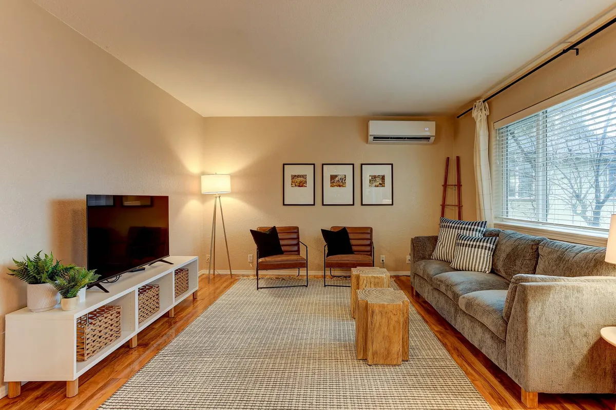 The image showcases a beautifully styled living room with a plush sofa, two inviting armchairs, a modern coffee table, a large wall-mounted television, tasteful artwork, and a warm floor lamp, all set in a cozy, sunlit space ideal for relaxation.
