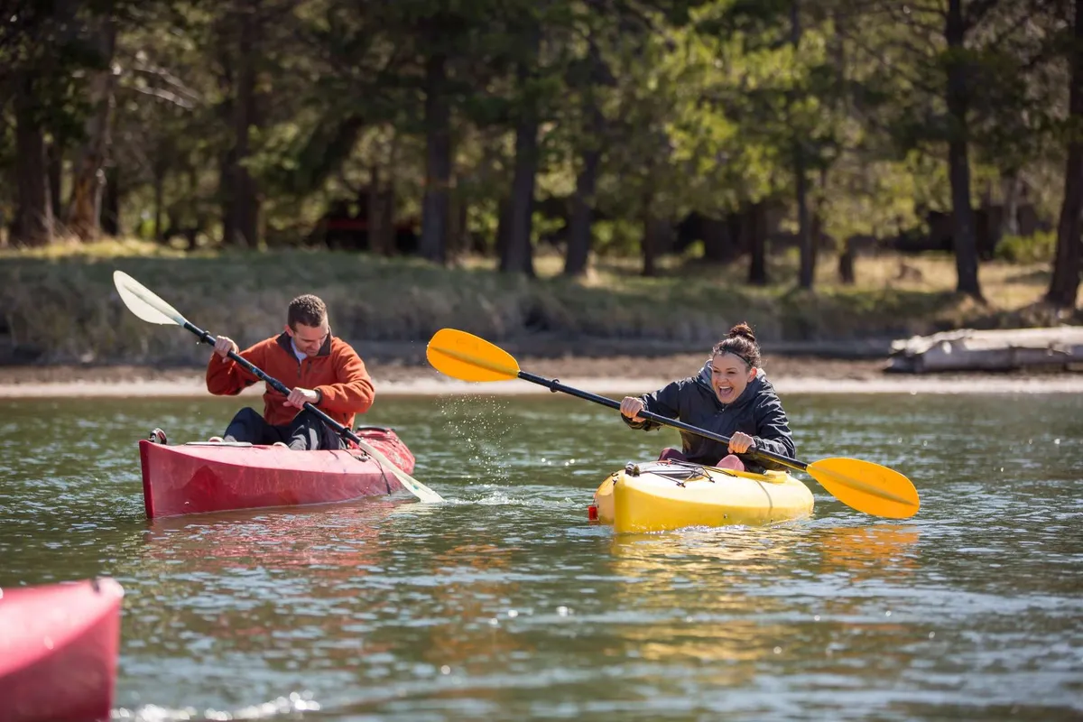 Two kayakers glide across calm water, one in a bright yellow kayak and the other in a red kayak. Reflected sunlight sparkles on the water’s surface, creating a dynamic outdoor scene rich with movement and adventure.