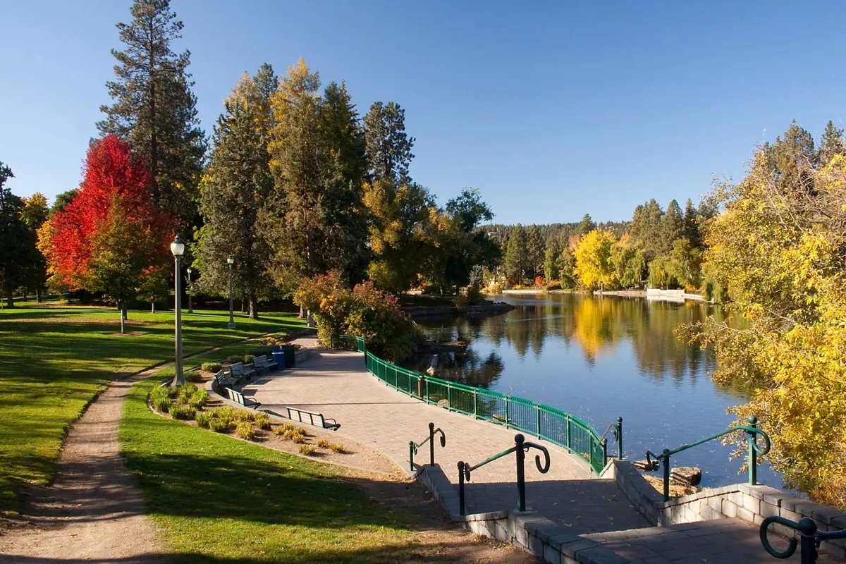 This exterior shot captures a tranquil lakeside park with a pathway winding among trees, benches, and a picturesque bridge. The setting suggests opportunities for walking, relaxing, and enjoying community events.