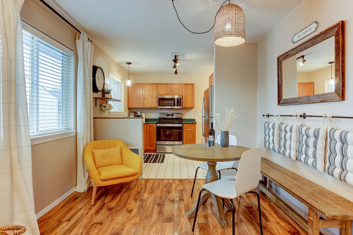A bright dining and kitchen area with a modern table, yellow accent chair, and a large window with curtains that frame views outside. The layout invites both casual dining and sociable gatherings.