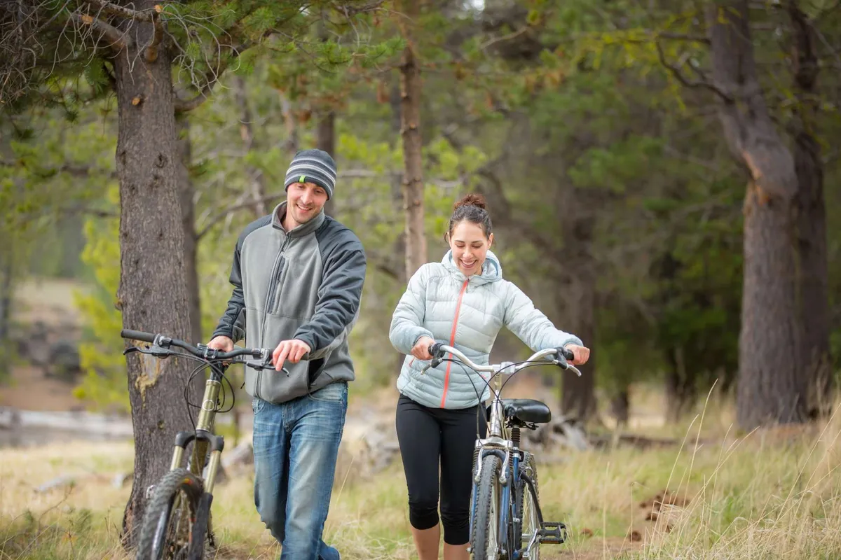 Two individuals walk with their bicycles through an open grassy area surrounded by tall trees, evoking the feel of an easygoing urban adventure. The mood is joyful and relaxed, highlighting outdoor recreation.