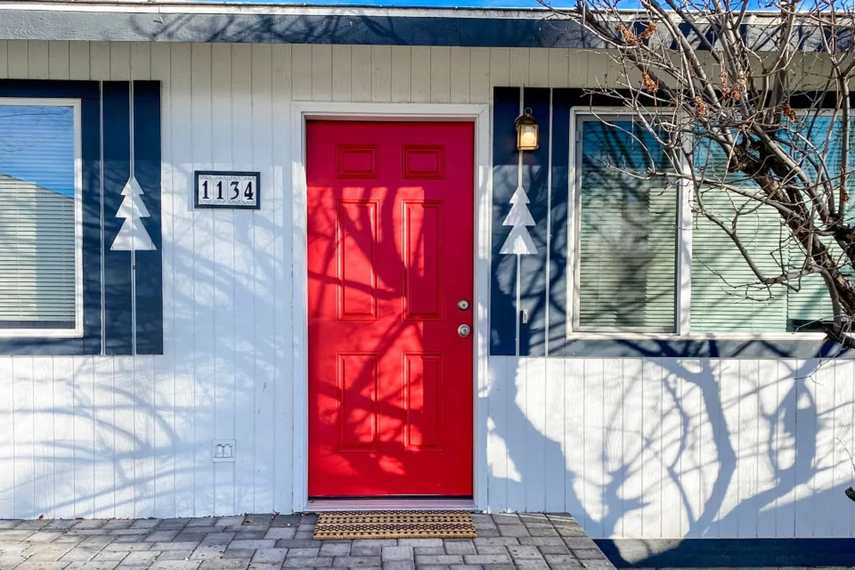 The image shows a cheerful red front door with a house number, flanked by decorative tree shapes and welcoming shadow patterns on the wall. The scene creates a sense of arrival and hints at the vibrant life just outside.