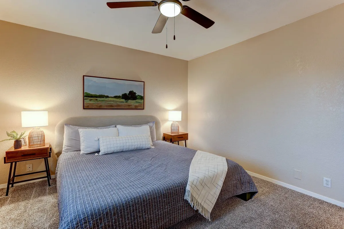 A serene bedroom with a bed featuring patterned bedding, flanked by nightstands with lamps and adorned with landscape artwork. The space feels peaceful and bright, ideal for relaxing, reading, or waking up gently.