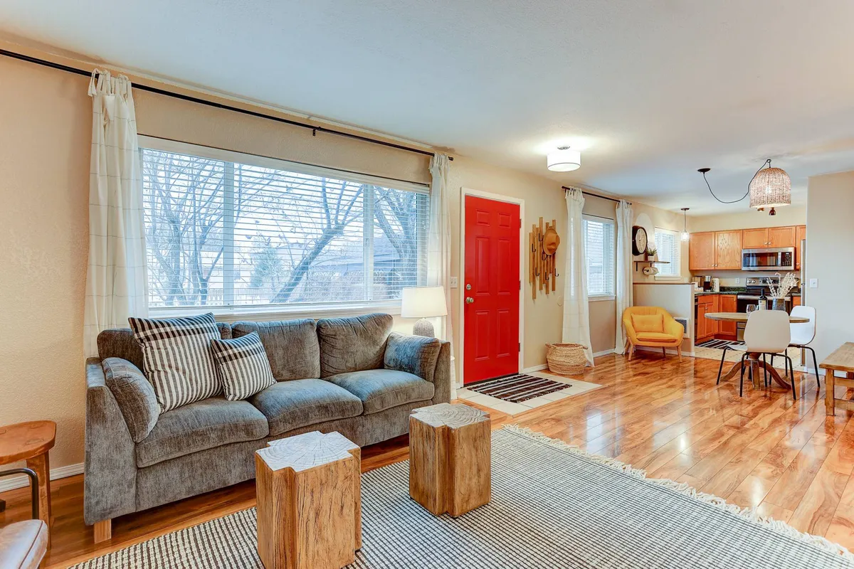 This image showcases an inviting open-concept space featuring a comfortable gray couch, wooden coffee tables, and a clear view of the adjacent kitchen area. The red front door and natural light contribute to a warm, communal ambiance where social gatherings and family meals can easily take place.