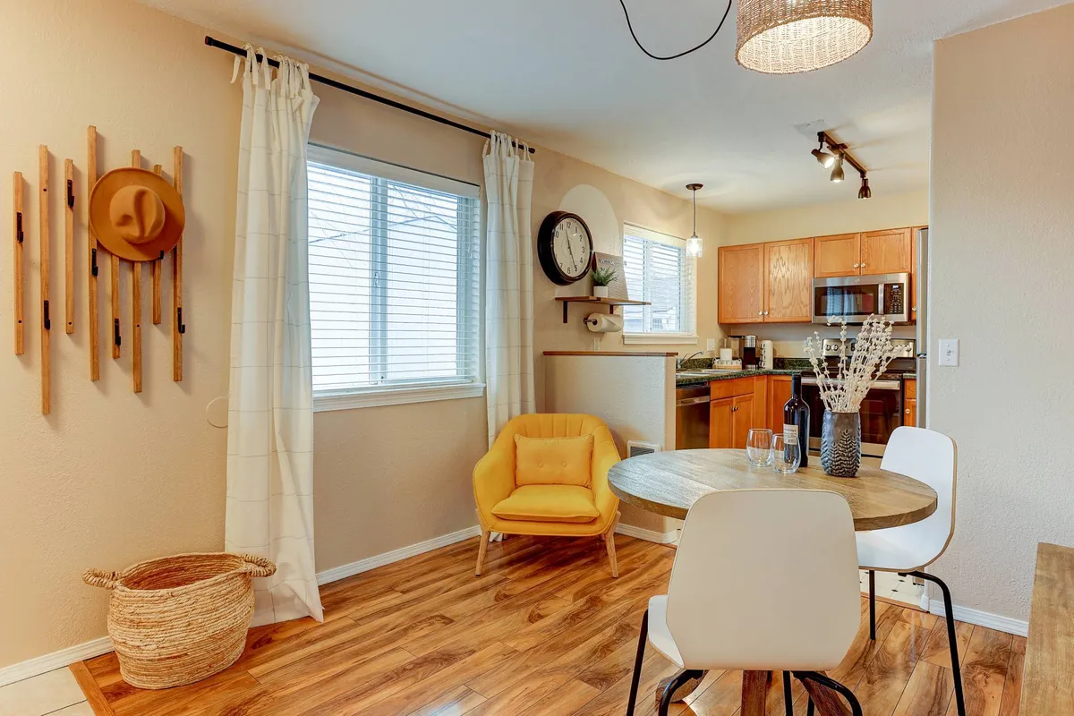 A cozy dining area featuring a wooden table with a vibrant yellow chair, a stylish hanging light fixture, wall hooks with decorative touches, and a large window that brings in natural light. The space is set up for enjoying meals, socializing, or relaxing with a book or work by the window.