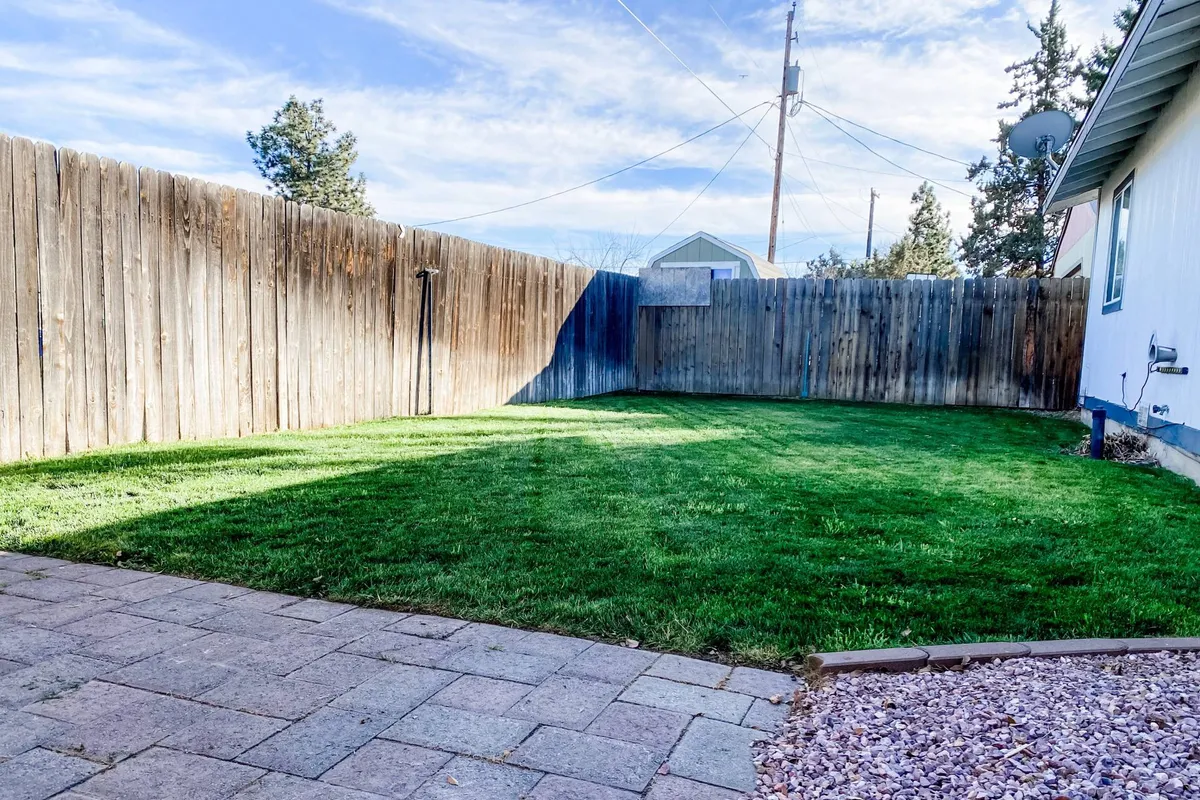 A view of a well-maintained lawn bordered by a wooden fence, accompanied by a paved patio area. The space is set up for outdoor gatherings, morning coffee on the patio, and relaxed evening stargazing, with clear indications of privacy and security.